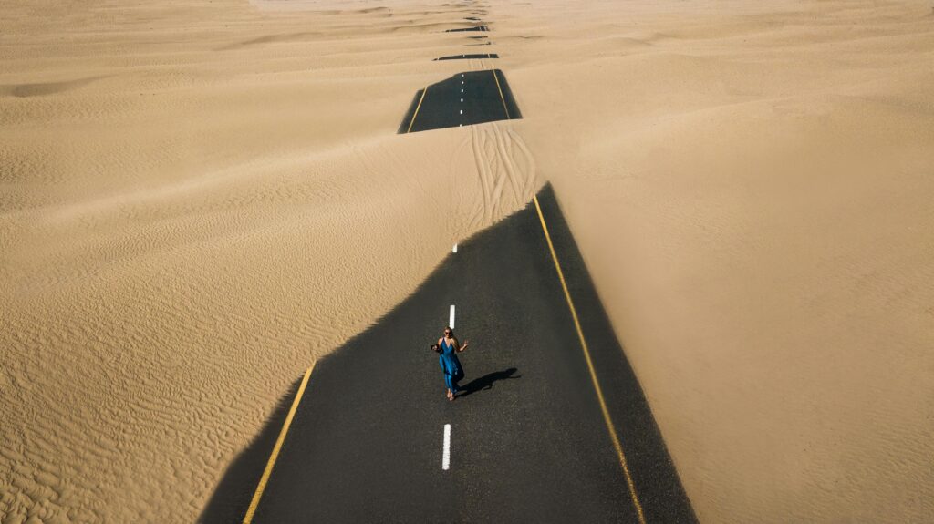 A lone woman walking on a sand-covered road in Dubai's desert, captured from above.