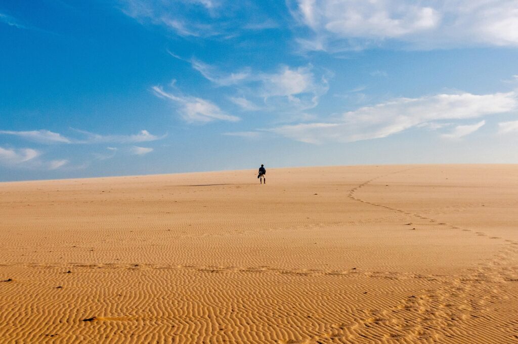 A lone traveler walks across a vast and serene desert under a bright blue sky.